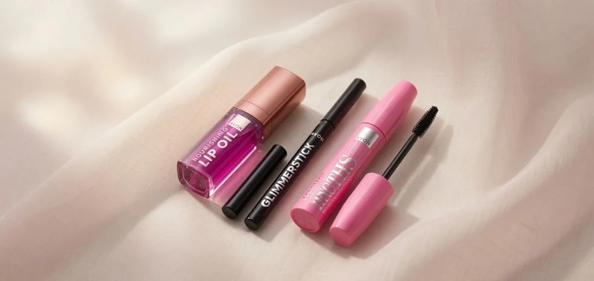Tři produkty AVON — lip oil, tužka Glimmerstick a řasenka Legendary Lengths — na jemné průsvitné látce v teplém minimalistickém stylu.